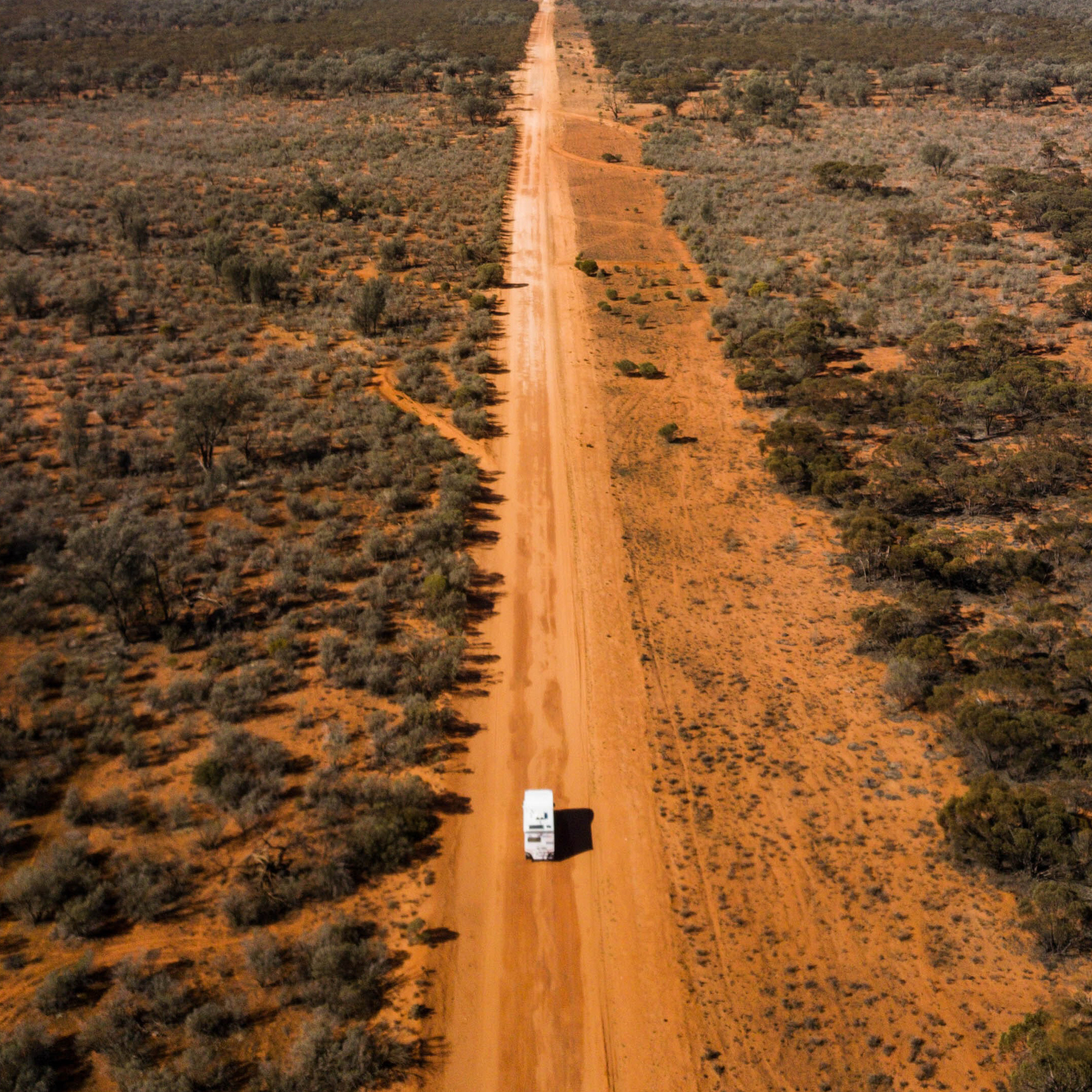An aerial view of a truck driving along a long, red, desert road.
